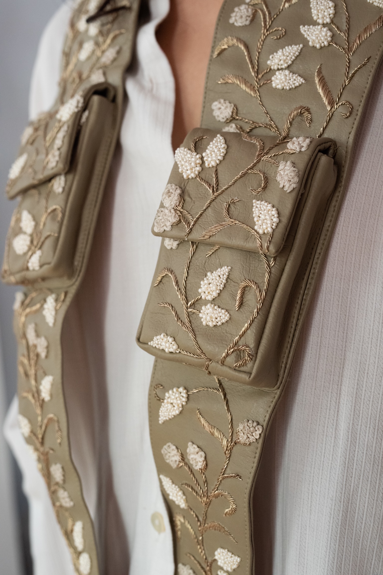 spero: olive leather straps with pockets, hand-embroidered with gilded leaves, worn over a white shirt